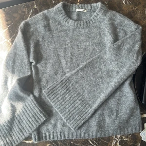 Grå stickad tröja från By Malene Birger - Säljer en stilren grå stickad tröja från By Malene Birger. Tröjan har en klassisk rund hals och långa ärmar med ribbade muddar. Perfekt för kyliga dagar!
