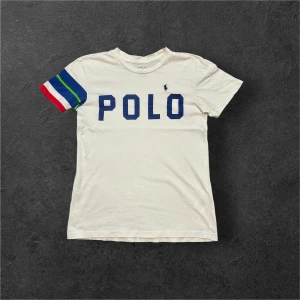 Vit t-shirt från Ralph Lauren - Säljer en vit t-shirt från Ralph Lauren med blå text 'POLO' på framsidan. Ena ärmen har färgglada ränder i blått, grönt, gult och rött. Perfekt för en avslappnad stil! Defekter som på bilden plus två små hål i kragen, skriv gärna för bild!
