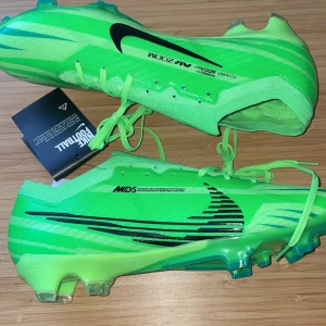Nike Mercurial grön  - Snygga gröna Nike fotbollsskor med svarta detaljer och snörning. Skorna har en modern design med Zoom-teknologi för optimal prestanda på planen. Perfekta för fotbollsspelare som vill ha både stil och funktion.