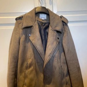 Brun mockajacka från Shine Original - Snygg brun mockajacka från Shine Original med dragkedja och knappar. Jackan har långa ärmar och en klassisk krage. Perfekt för höst och vår med sin stilrena design.