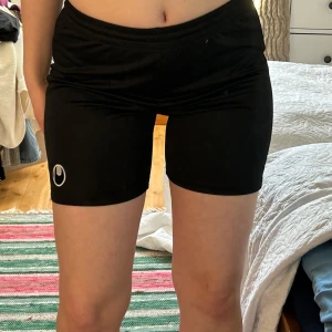 Svarta träningsshorts med logotyp - Säljer ett par svarta träningsshorts med elastisk midja och en liten logotyp på benet. Perfekta för gymmet eller löprundan. De är bekväma och ger bra rörelsefrihet. Passar S. 
