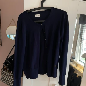 Mörkblå kofta från Holly & Whyte - Säljer en stilren mörkblå kofta från Holly & Whyte. Koftan har guldiga  knappar framtill och en klassisk rund halsringning. Är inte helt säker på storleken då den lappen inte längre sitter på tröjan men jag tor att det är storlek L men passar M också. 