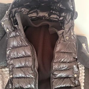 Moncler cardigan - Snygg svart cardigan från Moncler med quiltad design och dragkedja. Västen har en ribbad insida och praktiska fickor med knappar. Perfekt för kyligare dagar när du vill hålla stilen. Den har NFC tagg som går att blippa med mobilen och en qr kod att skanna via kamera. Perfekt för alla tider och säsonger.
