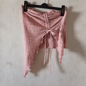 Rosa topp med snörning - Säljer en söt rosa topp med snörning framtill och en unik asymmetrisk design. Toppen har en texturerad yta och är perfekt för att ge en trendig touch till din outfit. Passar bra till både vardag och speciella tillfällen.