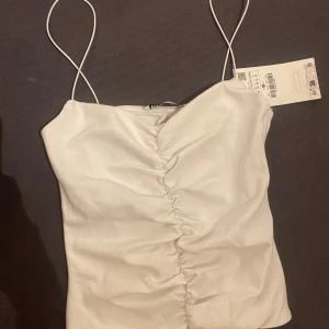 Vit linnetopp från Zara - Säljer en stilren vit linnetopp från Zara med smala axelband och rynkdetalj framtill.  (Liten i storlek )