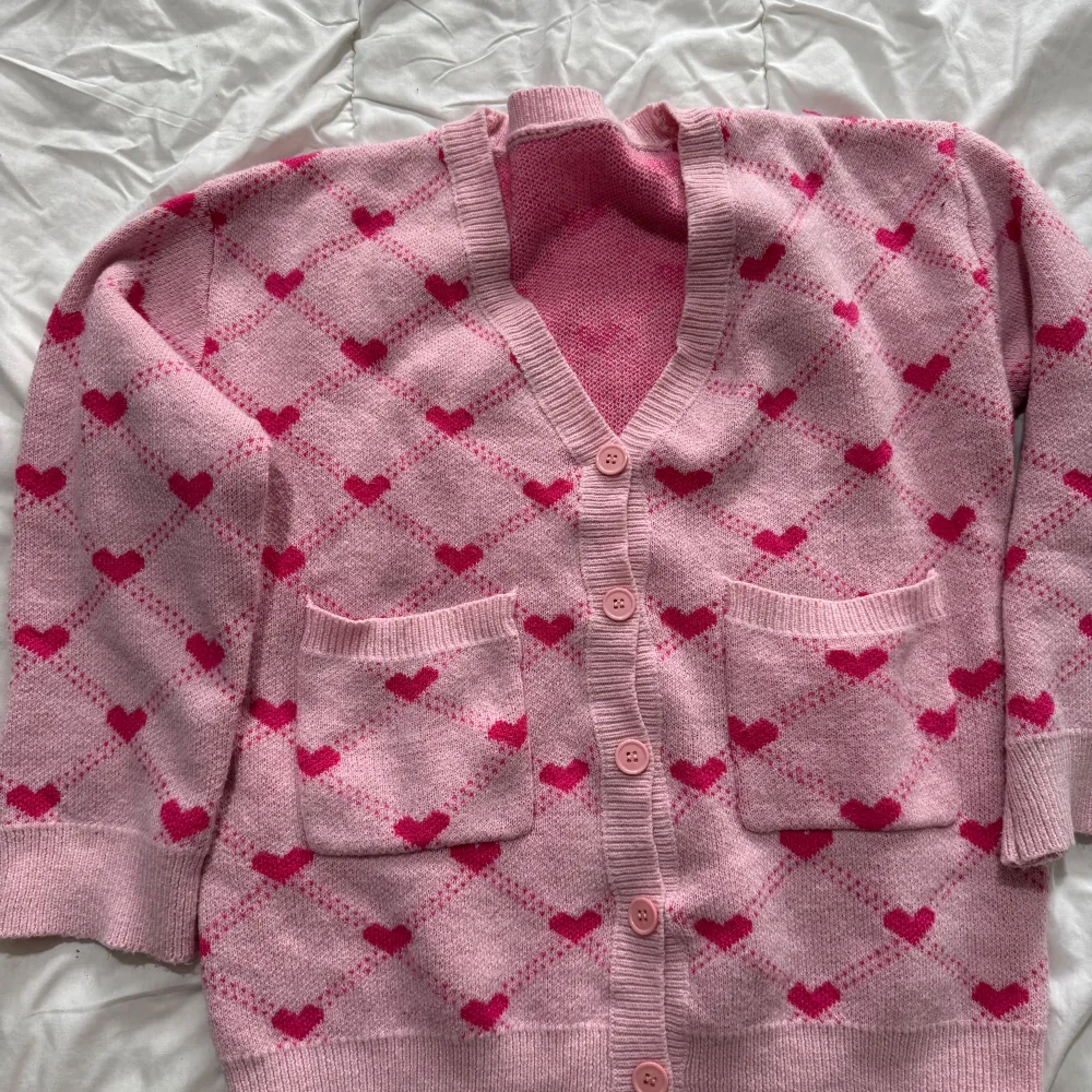 Söt rosa kofta med hjärtmönster i en ljusare nyans. Koftan har knappar framtill och två fickor. Perfekt för en mysig dag.. Neuletakit & Villapaidat.