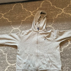 Grå hoodie från Polo Ralph Lauren - Säljer en stilren grå hoodie från Polo Ralph Lauren med dragkedja och klassisk logga på bröstet. Perfekt för en avslappnad look. Den har en bekväm passform och är tillverkad i mjukt material. Det är storlek L 14-16, 160. Det är inga defekter, tröjan är bara använd mycket men annars är det bra skick. Skriv om ni har några funderingar elr vill köpa😎