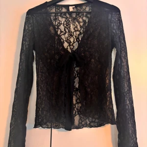 Svart spetsblus från Nelly - Säljer en elegant svart spetsblus från Nelly i storlek XS. Blusen har långa ärmar och en v-ringad design med knytning framtill. Perfekt för en stilfull look.