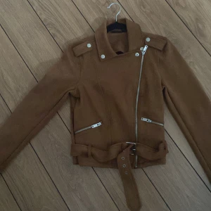 Brun jacka med bälte - Snygg brun jacka med dragkedja och bälte. Jackan har långa ärmar och en cool asymmetrisk design. Perfekt för höst och vår.