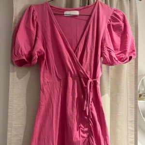 Rosa omlottklänning från Samsa Collection - Snygg rosa omlottklänning från Samsa Collection med puffärmar och volangdetaljer. Klänningen har en knytning i midjan och en v-ringad hals. Perfekt för en somrig look! 🌸använd 1 gång!🩷