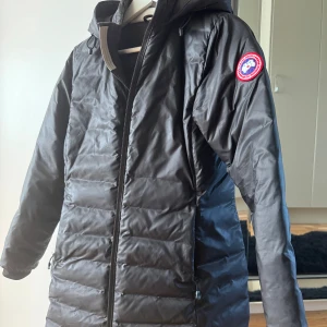 Svart dunjacka från Canada Goose - Säljer en stilren svart dunjacka från Canada Goose. Jackan har en lång passform med dragkedja och en ikonisk logga på ärmen. Perfekt för kyliga vårdagar med sin varma och lätta design.