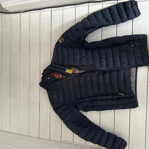 Mörkblå  lättviktsjacka från Parajumpers - Säljer en svart superlätt dunjacka från Parajumpers. Jackan har en dragkedja framtill och en snygg patch på ärmen. Perfekt för kyliga dagar med sin quiltade design och långa ärmar. Inuti är den grå med märkets logga och detaljer i orange.