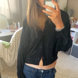 Svart ribbad kofta - Säljer en svart ribbad kofta med knappar framtill. Koftan har en croppad passform och långa ärmar. Perfekt för en stilren look. Köpt i Thailand 