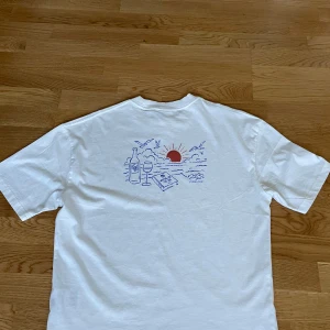 Vit t-shirt från Solid - Säljer en vit t-shirt från Solid med ett tryck av en solnedgång, vinflaska och glas på baksidan. Framsidan har en mindre version av samma tryck med texten 'Waves & Space'. Gjord av 100% bomull, perfekt för en avslappnad stil. Stolen S, passar även M. Använd 1 gång