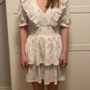 Vit blommig klänning från Gina Tricot - Charmig vit klänning med blommigt mönster från Gina Tricot. Den har volangdetaljer och en v-ringad hals. Perfekt för sommarens tillfällen.