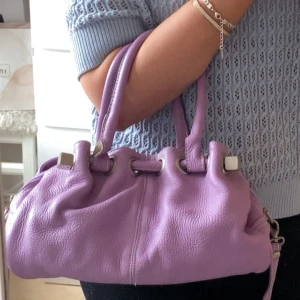 Lila handväska med dragkedja - Snygg lila handväska i skinnmaterial med dragkedja och handtag. Perfekt för att ge en färgklick till din outfit. Väskan har en elegant design med metalliska detaljer. Går två fläckar på insidan kan skicka bild vid behov. Medföljer också ett crossbody-strap