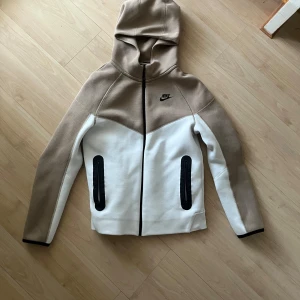 Beige och vit jacka från Nike - Säljer en snygg beige och vit zip hoodie från Nike med huva och dragkedja. Jackan har långa ärmar och svarta detaljer vid fickorna. Perfekt för en sportig look!