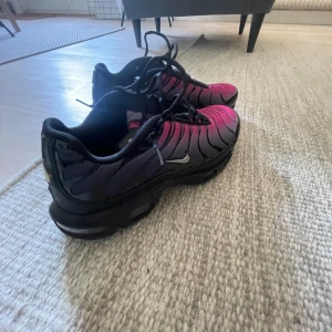 Nike tn rosa/svarta sneakers - Nike Tn sneakers med grymt snygg färgskiftning från rosa till svart och svarta detaljer. Skorna har klassisk Tn-logo på hälen, svarta snören och en vågig design på ovandelen. Sulan är svart och har synlig Air Max-dämpning. Perfekta för dig som vill sticka ut.