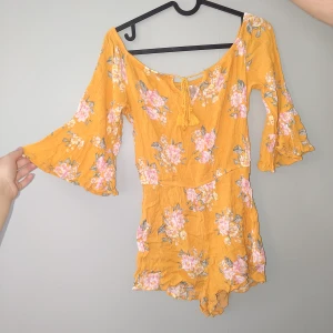 Blommig playsuit - Superfin gul playsuit med rosa blommor och volangärmar. Playsuiten har liten offshoulder-modell, knytning fram och är tillverkad i ett lätt, skönt material. Perfekt för sommarens alla tillfällen!