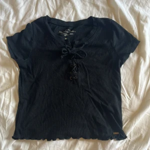 Svart ribbad snörningstopp Hollister - Svart croppad topp från Hollister med ribbad struktur och snörning framtill. Kortärmad modell med lätt vågig kant nertill och metallöljetter vid snörningen. Perfekt för dig som gillar en edgy och trendig stil.