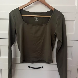 Olivgrön långärmad topp Hollister - Säljer en olivgrön långärmad topp från Hollister Must-Have Collection. Toppen har en snygg fyrkantig ringning och är figurnära i modellen. Perfekt att matcha med jeans eller kjol för en clean och stilren look.