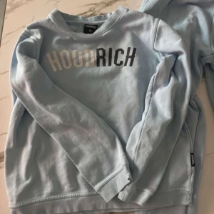 Ljusblå sweatshirt från Hoodrich  - Ljusblå sweatshirt från Hoodrich i storlek XL (13-15 år). Tröjan har rund hals, långa ärmar och tryckt HOODRICH-logga i vitt och svart framtill. Mjuk insida och ribbade muddar vid ärmslut och nederkant. Perfekt för chill dagar.