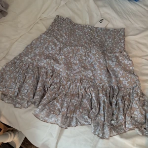 Brun o blå blommig volangkjol från lager 157 - Supersöt kort kjol från lager 157 i ljusbrunt med blått blommönster. Kjolen har bred smock i midjan och flera volanglager som ger en flowy känsla. Perfekt för dig som gillar romantisk och avslappnad stil. Prislapp kvar, aldrig använd.