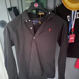 Svart skjorta från Ralph Lauren - Svart slim fit skjorta från Ralph Lauren i 100% bomull med stretch. Klassisk design med knappar framtill och rött broderat Polo-logo på bröstet. Långärmad och perfekt för en clean och stilren look. Skjortan är lite sol bränd men inte super mycket.