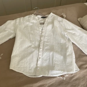 Vit broderad skjorta från Zara XS - Superfin vit skjorta från Zara i storlek XS med broderade detaljer och knappar framtill. Skjortan har lång ärm och är tillverkad i ett lätt bomullstyg, perfekt för vår och sommar. Snygg v-ringning och romantisk känsla tack vare spetsmönstret.