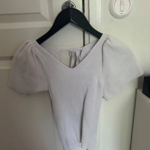 Vit ribbad topp med puffärm - Supersöt ljuslila ribbad topp med v-ringning och puffiga korta ärmar. Toppen har knytning i ryggen som ger en extra detalj och sitter snyggt på kroppen. Perfekt för dig som gillar en feminin och trendig stil.