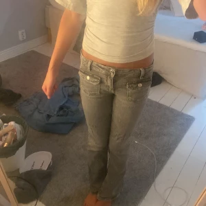 Gråa lågmidjade bootcut jeans - Säljer ett par gråa jeans med låg midja och bootcut-ben. Jeansen har snygga fickor framtill med lock och knappar, samt klassiska bakfickor. De är i ett mjukt denimtyg och har en riktigt chill och trendig vibe.