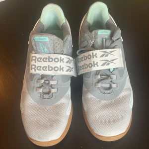 Grå Reebok Legacy Lifter träningsskor - Reebok Legacy Lifter träningsskor i grått med vita och mintgröna detaljer. Skorna har snörning och bred kardborre över vristen för extra stabilitet. Ovandelen är i syntet och mesh, sulan är robust och designad för lyft. Perfekta för gymmet och styrketräning.