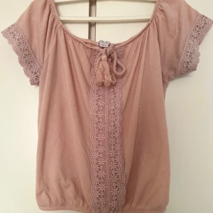 Rosa blus med spets från Amisu - Supersöt ljusrosa blus från Amisu med korta ärmar och bred resår nertill. Blusen har spetsdetaljer längs ärmslut och framtill, samt knytning med tofsar vid halsen. Perfekt för dig som gillar romantiska och boho-vibbar.