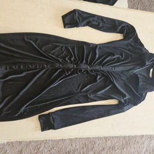 Svart långärmad klänning från Ellos - Svart långärmad klänning från Ellos i mjukt, glansigt material. Klänningen har knappar framtill, rynkade detaljer vid midjan och en smickrande passform. Perfekt för dig som gillar stilrena och bekväma plagg med en twist.