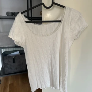 Vit ribbad blus med spetskant - Supersöt vit blus från Brandy Melville med ribbat tyg och korta ärmar. Urringningen är dekorerad med en fin spetskant som ger en romantisk vibe. Tyget är lätt och luftigt, perfekt för varmare dagar. Små hålmönster i tyget ger extra detalj. Passar till både jeans och kjol.