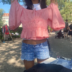 Rosa offshoulder blus med volang - Supersöt ljusrosa blus med offshoulder och puffiga trekvartsärmar. Toppen har smockad midja, volangkant nertill och liten knytning framtill. Perfekt för sommarens alla häng och picknickar. Luftig och lätt i bomull.