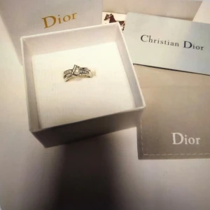 Dior silverring med stenar - Snygg ring från Dior i silverfärg med glittrande stenar och en unik, böjd design. Ringen har dubbla band som korsar varandra och är dekorerad med små, gnistrande stenar på ena sidan. Perfekt för dig som vill sticka ut med en lyxig accessoar.