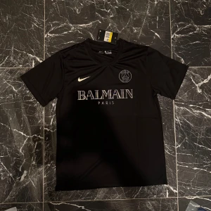 Svart PSG x Balmain fotbollströja Nike S - Snygg svart fotbollströja från Nike i samarbete med Paris Saint-Germain och Balmain. Tröjan har korta ärmar, rund hals och tryckta loggor på bröstet. Materialet är lätt och andas, perfekt för träning eller match. Stilsäker design med exklusiv känsla.