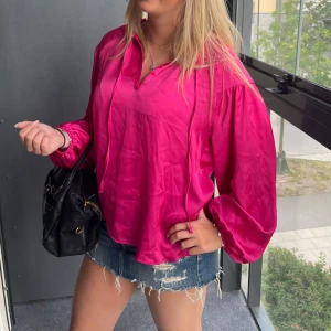 Ceriserosa blus i sidenlook - Säljer en ceriserosa blus med ballongärmar och v-ringning. Blusen har knytband vid halsen och är tillverkad i ett glansigt material som ger en lyxig känsla. Perfekt för dig som vill sticka ut med färg och stil.