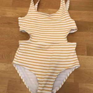 Randig baddräkt från Zara i gult och vitt - Superfin baddräkt från Zara med gula och vita ränder. Modellen har coola cut-outs i midjan och breda axelband. Materialet är ribbat och stretchigt, perfekt för strand eller pool. Snygg och bekväm för sommardagar!