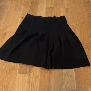 Svart plisserad kortkjol med dragkedja - Svart kortkjol med plisserade veck och hög midja. Kjolen har dragkedja i sidan och knappstängning i midjan, samt justerbar resår på insidan för perfekt passform. Enkel och stilren design som är lätt att matcha.