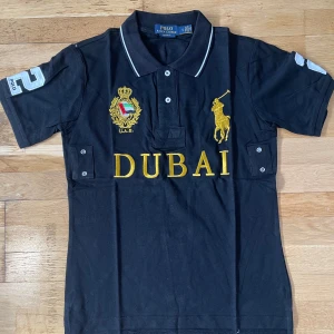 Svart pikétröja Polo Ralph Lauren S - Svart pikétröja från Polo Ralph Lauren med broderad gul logga, DUBAI-text och emblem framtill. Stora vita siffror på rygg och ärm, klassisk krage med vita detaljer och knappar. Tillverkad i bomull och har korta ärmar. Snygg och sportig vibe.