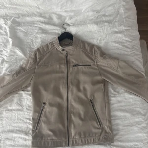 Beige skinnjacka med dragkedjor - Snygg beige skinnjacka med raka linjer och cool ståkrage. Jackan har tre dragkedjefickor framtill och stängs med dragkedja. Perfekt för dig som gillar stilren och clean look.