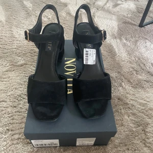 Svarta sandaler med klack från Novita - Stilrena svarta sandaler från Novita med bred klack och öppen tå. Skorna har en justerbar rem med guldfärgat spänne och är tillverkade i mocka. Använda en kväll och upptäckte de var för stora. Felfria. Nypris 999