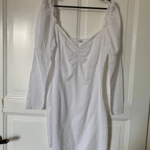 Vit kortklänning med puffärm - Supersöt vit kortklänning med långa puffärmar och lätt rynk vid bysten. Klänningen har subtila vertikala ränder i tyget och en romantisk v-ringad urringning. Perfekt för dig som gillar en feminin och trendig stil. Köparen betalar frakten 