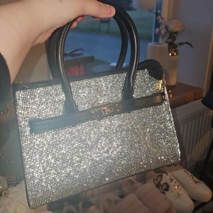 Glittrig silver handväska med svarta detaljer - Säljer en glittrig handväska i silver med svarta handtag och detaljer. Väskan har en klassisk fyrkantig form och stängs med ett snyggt spänne i metall. Perfekt för dig som vill sticka ut och addera lite extra bling till din outfit.