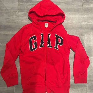 Röd hoodiejacka från GAP Kids XXL - Röd hoodiejacka från GAP Kids i storlek XXL. Skulle säga att den sitter som en xs i damstorlek och passar mig som är 174! Jackan har dragkedja framtill, huva och stora fickor. Klassisk GAP-logga i vitt och svart på bröstet. 