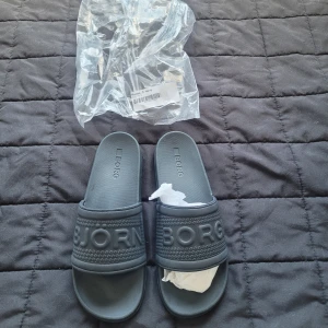 Mörkgrå slides från Björn Borg - Snygga mörkgrå slides från Björn Borg med tydlig logga över foten. Tillverkade i syntetmaterial med strukturerad rem och platt sula. Perfekta för sommaren eller till stranden.