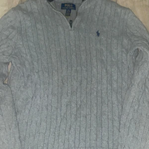 Polotröja - Kabelstickad grå tröja f med half zip och klassisk blå logga broderad på bröstet. Tröjan har ribbade muddar och krage med mörkblå ränder. Perfekt för lager-på-lager och snygg till jeans.