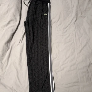 Svarta mönstrade mjukisbyxor Lacoste XL - Svarta mjukisbyxor från Lacoste i storlek XL med diskret geometriskt mönster, vita ränder längs sidorna och klassisk krokodillogga. Byxorna har elastisk midja med snörning och är tillverkade i mjukt material för maximal komfort.